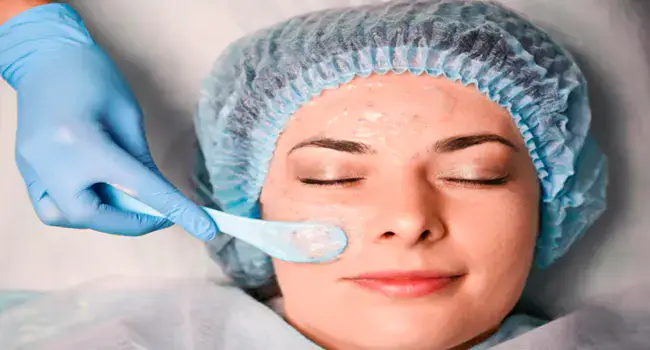 ممارسة بقفازات زرقاء تطبّق جلّاً شفافاً على خد مريضة بملعقة أثناء تقشير كيميائي في بيئة سريرية