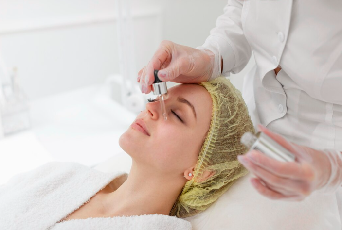Practitioner in a white coat applying hyaluronic acid skin booster serum with a glass dropper to a patient's cheek in a clinical setting in Dubai