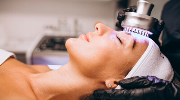 Professional light-based melasma treatment with a handheld device emitting purple light being applied to a patient's forehead by a practitioner in black gloves in a clinical setting
