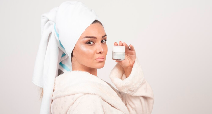 A woman in a white bathrobe with a towel-wrapped head holding a professional skincare product jar, representing medical-grade skincare at Dr. Elham's clinic in Dubai