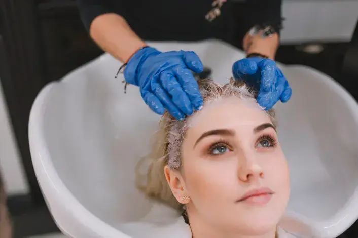 Practitioner in blue gloves applying a therapeutic scalp treatment to a woman's blonde hair during non-surgical hair restoration in Dubai