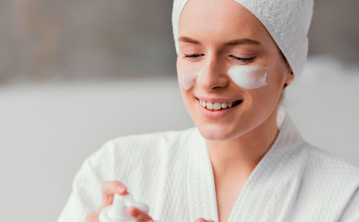 Close-up of a smiling woman wearing a white robe and head towel, with patches of white foam cleanser on her cheeks, as she dispenses product from a white pump bottle during a professional medical skincare routine in Dubai