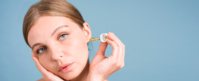 Young woman with clear skin holding a golden serum dropper against a soft light blue background, representing skin booster hydration treatment in Dubai