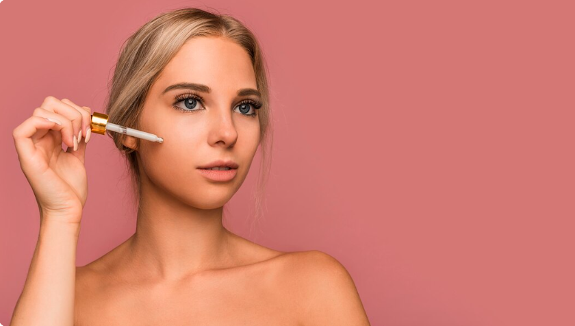 Young woman with clear skin applying golden skin booster serum with a glass dropper against a soft pink background, representing skin hydration treatment in Dubai