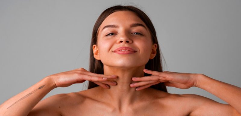 Smiling young woman with long dark hair gently touching her neck and jawline to highlight smooth firm skin against a neutral gray background, representing non-surgical skin tightening treatment in Dubai