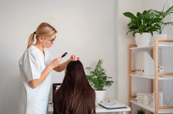 Specialist with glasses examining a patient's scalp with a magnifying glass beside a trichoscopy monitor to assess hair restoration candidacy in Dubai