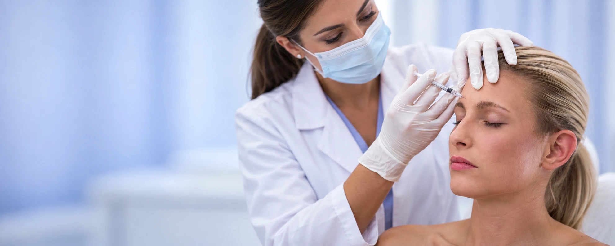 Female doctor in white coat and blue mask administering a forehead Botox injection to a blonde patient, wide clinical composition with soft blue copy space, representing professional Botox wrinkle treatment in Dubai
