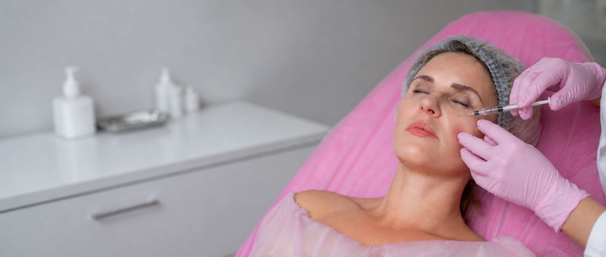 Practitioner in pink gloves administering a precise cheek and under-eye dermal filler injection to a patient in a surgical hair net on a pink treatment bed in a bright clinical setting, representing professional dermal fillers treatment in Dubai - Dr. Elham Keikha
