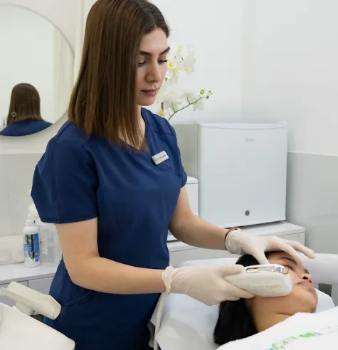أخصائية تجميل بزي أزرق تجري علاجاً لجلد الوجه بجهاز يدوي على خد مريضة في بيئة سريرية