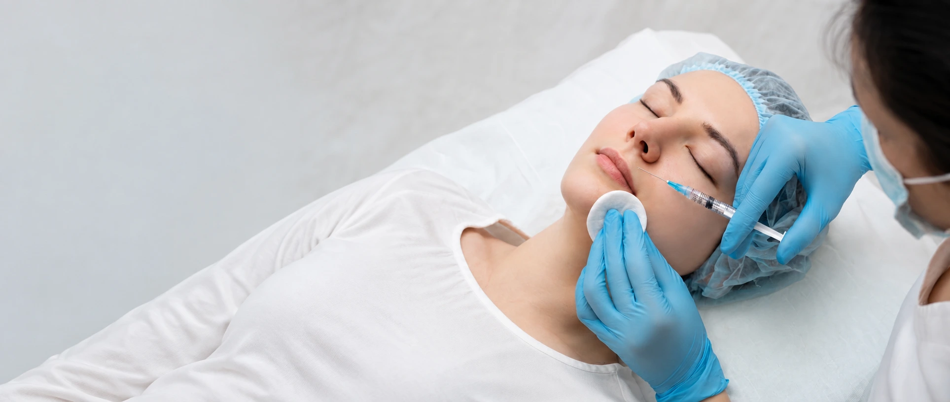 Clinical skin booster injection into the cheek of a relaxed patient wearing a surgical cap, performed by a gloved practitioner in a bright sterile clinic setting in Dubai - Dr. Elham Keikha